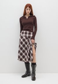 Top marrone a maniche lunghe con dettaglio zipper, abbinato a una gonna a quadri con uno spacco alto sulla coscia. Stivali neri al ginocchio con fibbia.