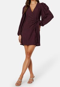 Vestido burgundy con mangas largas abullonadas y textura de rayas verticales, con un detalle de lazo en la cintura, combinado con tacones con tiras.
