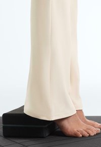 Pieds reposant sur un bloc de yoga noir, avec un pantalon large couleur crème couvrant les chevilles, debout sur un tapis noir texturé.