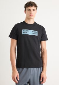 Juodas medvilninis marškinėliai su centre esančia šviesiai mėlyna grafika, kurioje užrašyta "CALVIN KLEIN NEW YORK". Lygus paviršius ir klasikinis apykaklės dizainas.