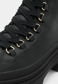 Bota de cuero negra con ojales de metal dorado, con cordones negros texturizados y una gruesa suela de goma negra. Acabado suave con detalles de costura.