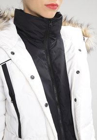 Osoba v bílém péřovém kabátu s kapucí lemovanou kožešinou, pod ním má zapnutou černou vnitřní bundu s vysokým límcem, viditelné rty s červeným leskem na rty.