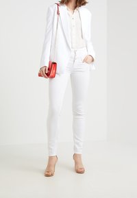 Vit blazer över en ljus skjorta, kombinerad med vita skinny jeans och beige klackar. En liten röd handväska med kedjes snodd ger kontrast.