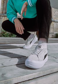 Baskets en suède gris avec une semelle blanche texturée, des accents noirs et des lacets ; branding visible sur la languette. Stylisées avec un pantalon noir et des chaussettes.
