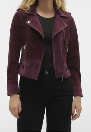 Mujer vistiendo una chaqueta biker de ante morado oscuro con detalles de cremallera sobre una camiseta negra y pantalones negros, de pie frente a un fondo liso.