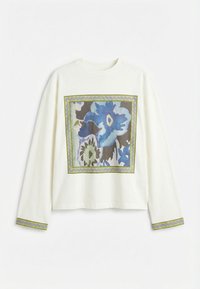 Λευκό πουκάμισο με μακρύ μανίκι, με μεγάλο μπλε τετράγωνο σχέδιο με λουλούδια στο εμπρόσθιο μέρος και αντίστοιχες λεπτομέρειες περιγράμματος στις μανσέτες των μανικιών.