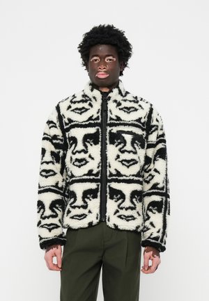 Fuzzy black and white jacket featuring repeated face pattern, zip closure, high collar, and textured fabric. Worn with dark green pants.