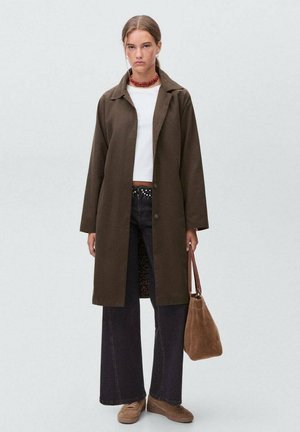 Giovane donna con un lungo cappotto marrone, top bianco corto, pantaloni neri a gamba larga, scarpe marroni, collana rossa e con una borsa marrone in mano.