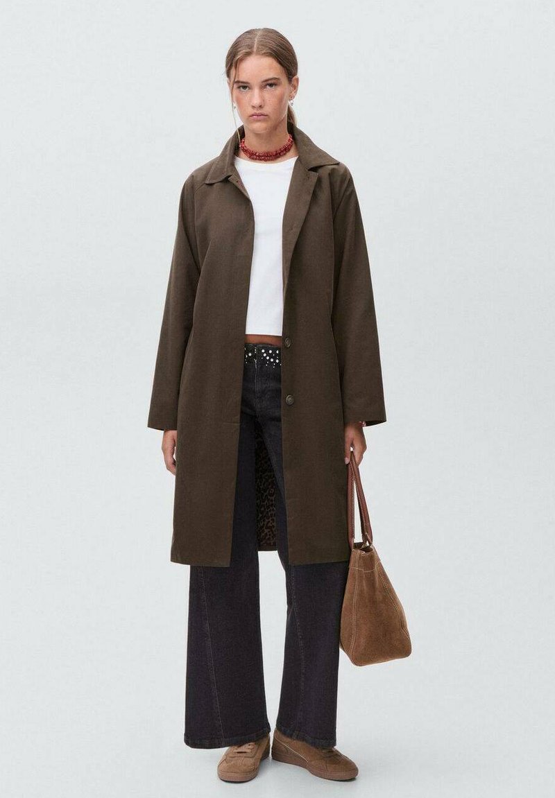 Joven mujer vistiendo un abrigo largo marrón, un top corto blanco, pantalones anchos negros, zapatos marrones, collar rojo y sosteniendo un bolso marrón.