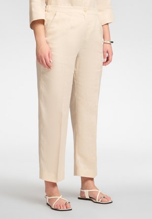 Beige hooggetailleerde cropped broek gedragen met beige platte sandalen met bandjes en een bijpassende top met lange mouwen, handen ontspannen langs de zijkant.