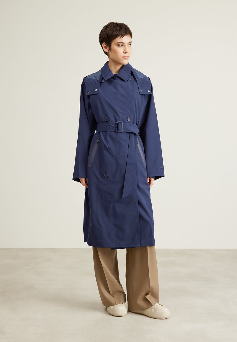 Manteau trench marine avec une ceinture, design croisé, épaulettes et poches latérales ; associé à un pantalon ample marron clair.