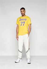 Κίτρινο μπλουζάκι με τη λέξη "Lakers" και τον αριθμό "77" σε μωβ χρώμα. Λευκό παντελόνι φόρμας με λεπτομέρειες από δίχτυ και γκρι αποχρώσεις. Λευκά αθλητικά παπούτσια.