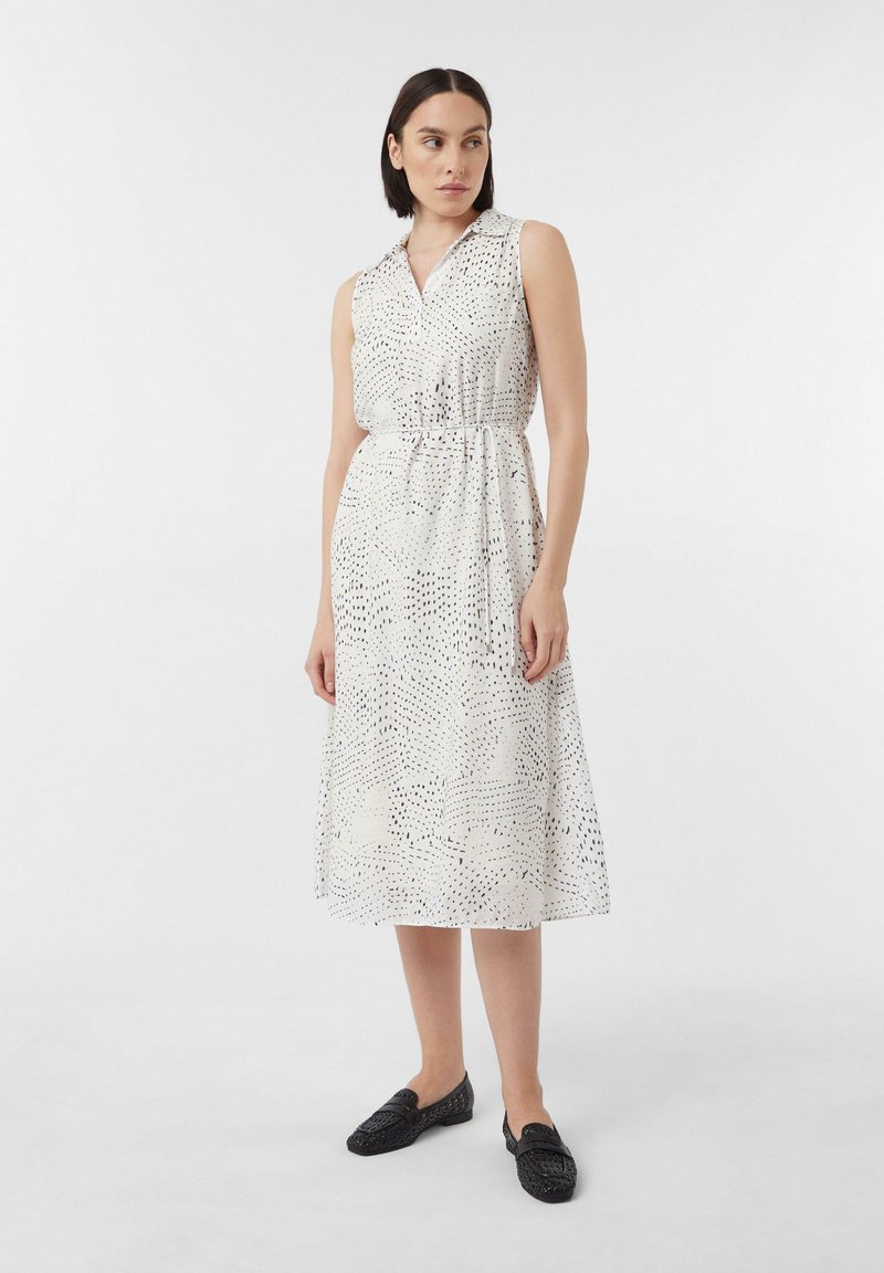 Ärmelloses weißes Kleid mit einem Kragen, das ein schwarzes Punktmuster und einen Taillengürtel aufweist, kombiniert mit schwarzen geflochteten Loafers.