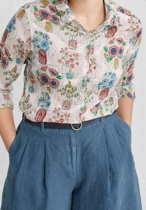Chemise fleurie boutonnée avec des tournesols et d'autres fleurs en vases, associée à un pantalon bleu plissé taille haute et une ceinture noire avec une boucle ronde.