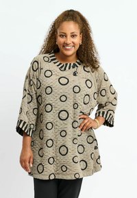 Beige Tunika mit schwarzen kreisförmigen Mustern und strukturiertem Finish. Verfügt über einen gestreiften Kragen und drei Viertel lange Ärmel mit Knopfdetails.