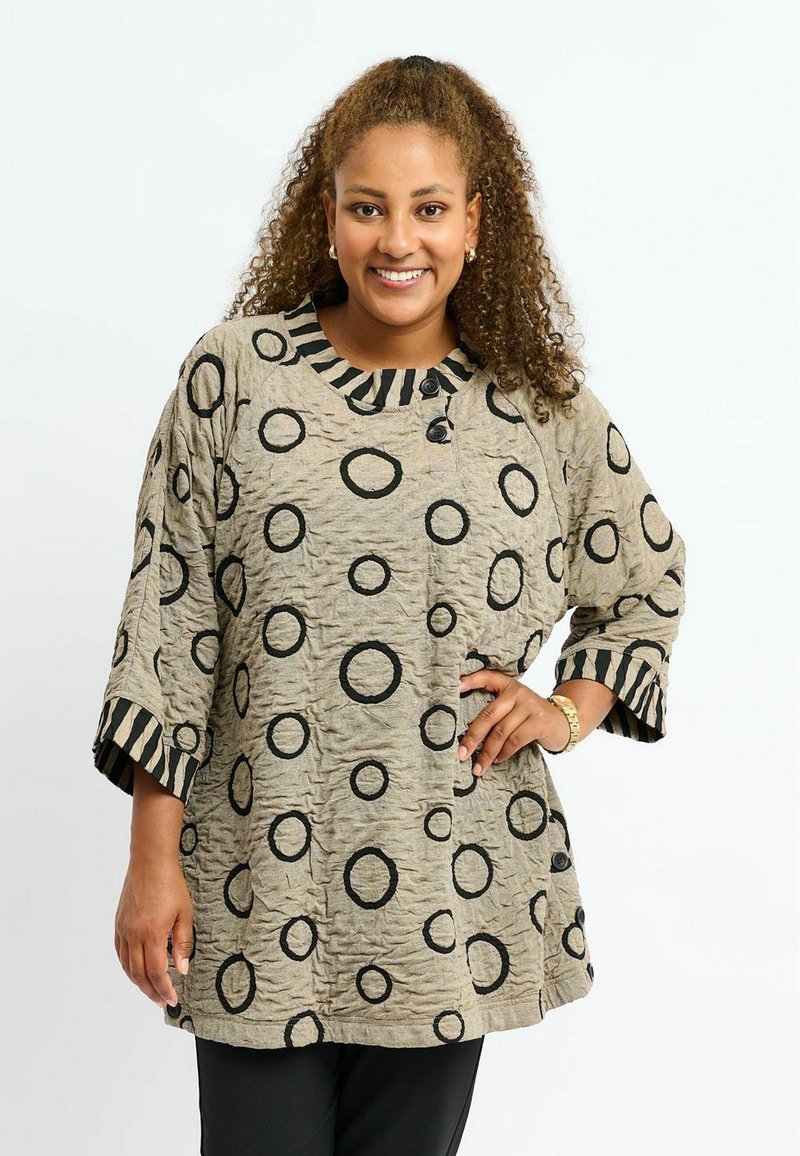 Beige Tunika mit schwarzen kreisförmigen Mustern und strukturiertem Finish. Verfügt über einen gestreiften Kragen und drei Viertel lange Ärmel mit Knopfdetails.