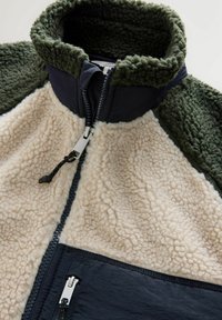 Fleece jakke med høj krave, der har beige shearling, mørkegrønne accenter og marineblå paneler. Lynlås lukning med en metallisk træk.