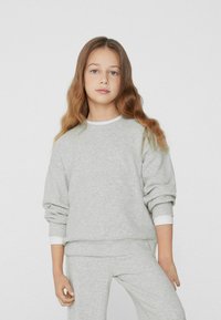 Světle šedá mikina vyrobená z měkkého materiálu, s bílým kulatým výstřihem a manžetami. Model pózuje s odpovídajícími kalhotami, zobrazuje uvolněný střih.