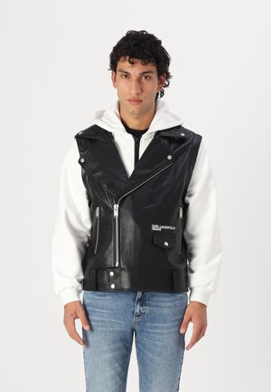 Black leather sleeveless biker vest with silver zippers and snaps, featuring front pockets and white hooded sweatshirt underneath. Denim jeans.