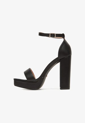 Czarna sandałka na wysokim obcasie z paskiem wokół kostki, masywną platformą, odkrytymi palcami, wykonana z gładkiej skóry o polerowanym wykończeniu.