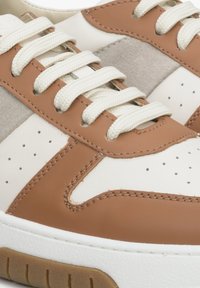 Białe skórzane sneakersy z brązowymi akcentami, zamszowym panelem bocznym, perforowanym czubkiem oraz płaskimi kremowymi sznurowadłami. Gruba gumowa podeszwa z brązowym bieżnikiem.