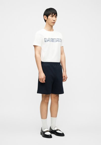 Νεαρός άνδρας στέκεται φορώντας λευκό μπλουζάκι με σχέδιο, ναυτικού χρώματος σορτς, λευκές κάλτσες και μαύρα και λευκά μοκασίνια μπροστά σε απλό φόντο.