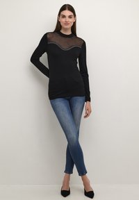 Schwarzes Langarmoberteil mit transparentem Mesh-Einsatz und geschwungenen Strassdetails; kombiniert mit blauen Skinny-Jeans und schwarzen spitzen Absatzschuhen.