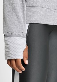 Close-up van een hand die een grijze top met gestructureerde mouwzoom en zwarte legging met witte zijstreep draagt.