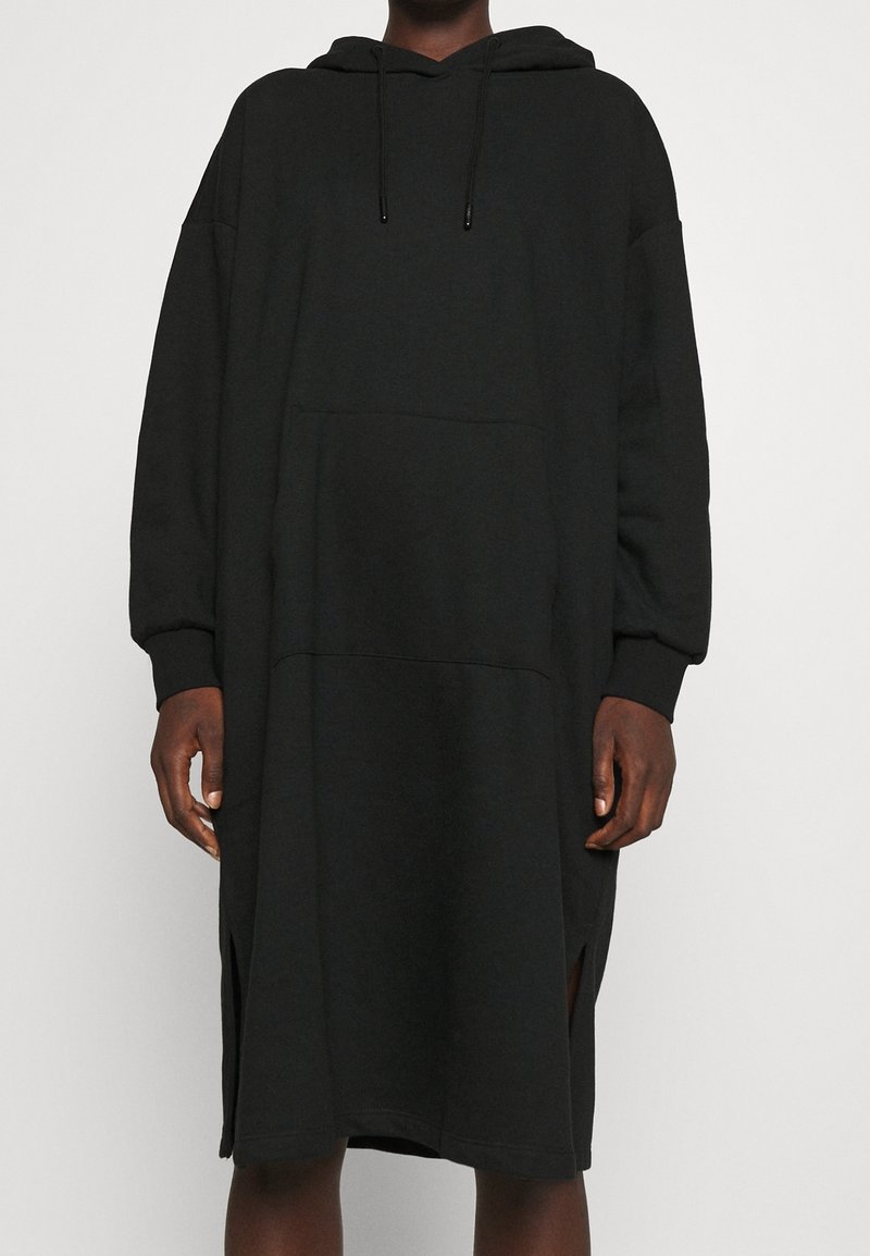 Personne portant une longue robe sweat-shirt noire ample à capuche avec poche avant et fentes latérales, debout devant un fond uni.