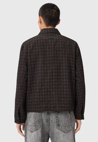 Veste à carreaux marron avec un col, réalisée en tissu texturé. Dotée de manches longues et d'une étiquette à l'arrière. Portée avec un pantalon en denim gris.