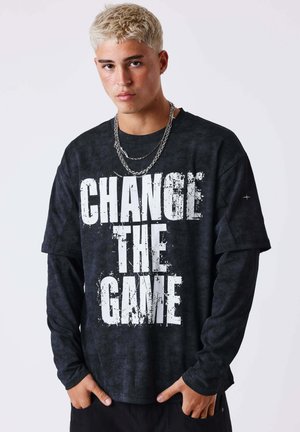 Joven con cabello corto y rubio que lleva varias capas de camisetas negras con el texto blanco "CAMBIA EL JUEGO" y dos collares de cadena plateada.