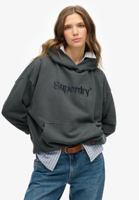 Felpa grigia oversize con tasca a marsupio e logo "Superdry" ricamato. Abbinata a una camicia a righe blu e jeans in denim.