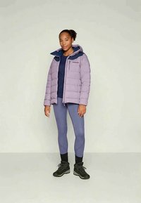 Moteris stovi apsirengusi šviesiai purpurine pūkine striuke, tamsiai mėlynu marškinėliais, purpurinėmis legginsais, juodomis kojinėmis ir juodais žygio batais ant vienspalvio fono.