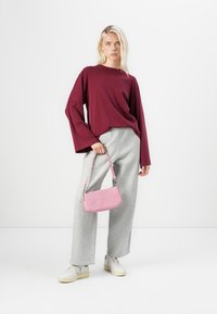 Bordeauxfarbene Langarmbluse, graue weit geschnittene Hose und weiße Turnschuhe; hält eine kleine, pinkfarbene, strukturierte Handtasche.