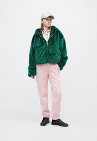 Osoba w zielonym, puszystym hoodie na zamek, jasnoróżowych dresach, czarnych trampkach, beżowej koszuli, szarym kapeluszu i złotych kolczykach w kształcie kół, stojąca na białym tle.