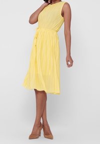Gelbes, plissiertes Midikleid mit ärmellosem Design und rundem Ausschnitt; beinhaltet einen Bindegürtel. Glatte Textur und fließender Stoff, kombiniert mit beigen Absätzen.