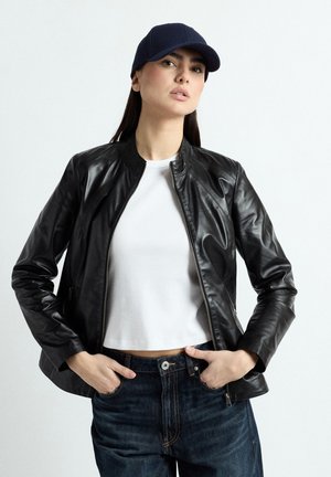 Kvinde iført en sort læderjakke, hvid cropped t-shirt, mørke jeans og mørkeblå baseballkasket, stående med hænderne i lommerne.
