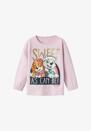 Világos rózsaszín, hosszú ujjú ing, amelyen mese kutyák láthatók, arany csillámos szöveggrafikával, ami azt írja: "ÉDES, AHOGY CSAK LEHET!" sötét háttérrel.