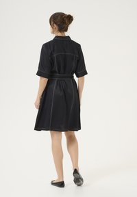 Melnā kleita ar īsām piedurknēm un jostu viduklī. Ar kontrastējošu baltu šuvumu un apkakli. A-veida forma ar ceļgaliem līdz sasniedzamu svārku iedobumu.