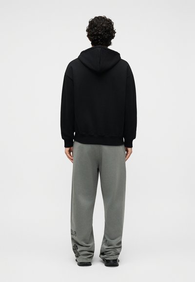 Άτομο με σγουρά μαλλιά που φοράει μαύρο φούτερ με κουκούλα και φαρδιά γκρι φόρμα με μαύρα αθλητικά παπούτσια, στέκεται γυρισμένο με την πλάτη σε αφηρημένο φόντο.