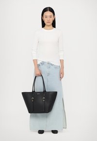 Sac en cuir noir avec un large fond et un sommet rétréci, doté de quincaillerie dorée, de poignées doubles et d'une texture lisse.