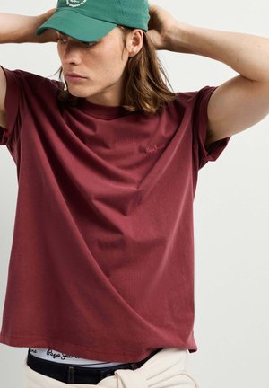Giovane con capelli fino alle spalle che indossa un berretto verde e una maglietta bordeaux larga, con le mani dietro la testa.