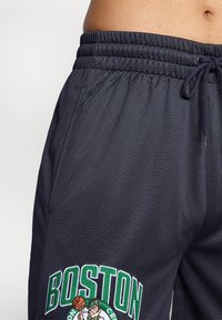 Sorte atletiske shorts med en struktureret overflade, elastisk talje og grønt broderet "BOSTON" logo med en leprechaun, der holder en basketball.