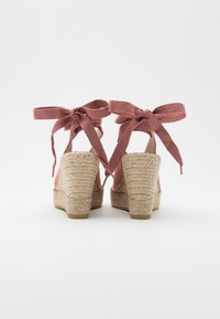 Pár béžových espadrilových klínových podpatků s látkovými kotníkovými vázáními v jemně růžové barvě, pohled zezadu na bílém pozadí.