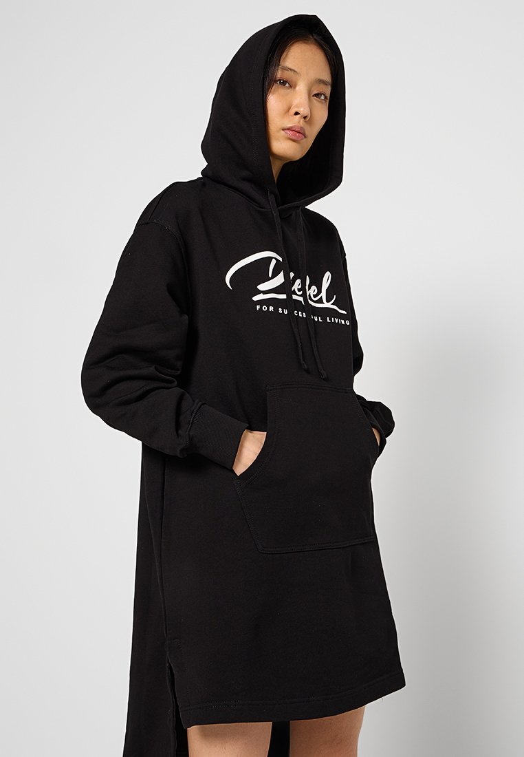Vrouw in een zwart oversized hoodie-jurk met een voorzak en witte tekst op de borst, staand tegen een effen lichte achtergrond.