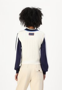 Knitted sleeveless sweater in cream with ribbed texture, blue striped collar, and a red logo patch at the back; paired with beige pants.