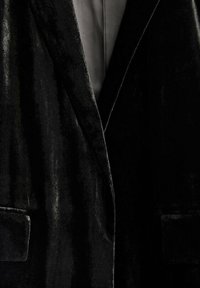 Blazer en velours noir avec col châle, deux poches avant et une doublure intérieure lisse, présentant une texture douce et un design classique.