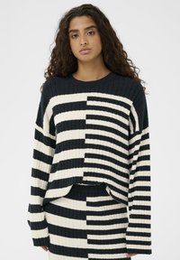 Gestrickter Pullover mit einer gerippten Textur, der ein gestreiftes Muster in Schwarz und Creme aufweist, mit entspanntem Schnitt und überschnittenen Schultern.