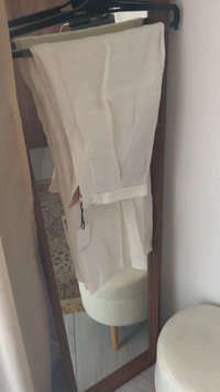 Biała lniana odzież wisząca na czarnym wieszaku, z przewiązanym pasem, krótkim rękawem i widoczną metką na materiale.