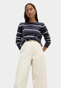 Sweter w marynarskie paski z białymi poziomymi liniami. Noszony z beżowymi spodniami o wysokim stanie i szerokimi nogawkami oraz czarnym paskiem. Gładka tekstura materiału.